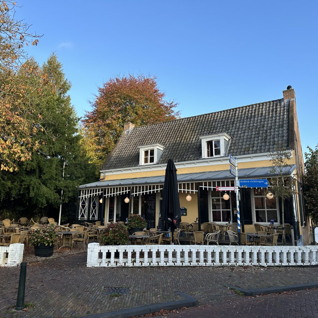Geel café-restaurant met wit hek en een terras vol lege rotanstoelen, omringd door herfstbomen.