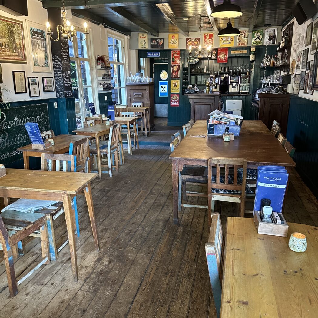 Binnenkant van een gezellig eetcafé met houten vloer, tafels, stoelen, een bar en de Buitenlust menukaart.