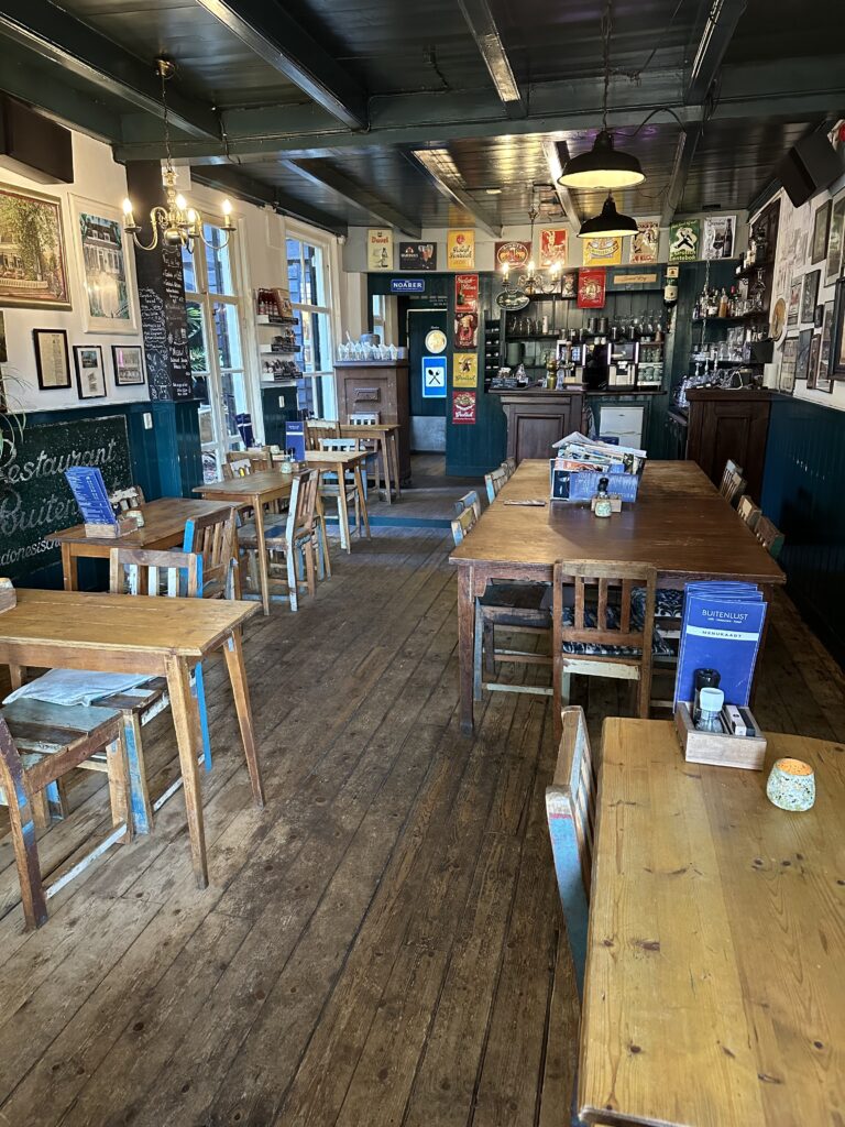Binnenkant van een gezellig eetcafé met houten vloer, tafels, stoelen, een bar en de Buitenlust menukaart.