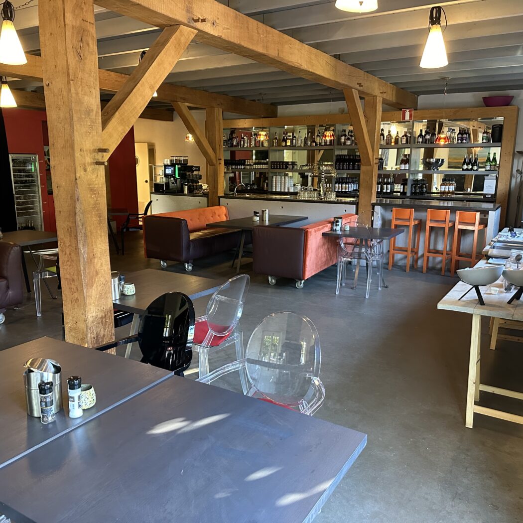 Modern café-restaurant interieur met houten balken, donkere tafels, banken, transparante stoelen en een bar met oranje krukken.