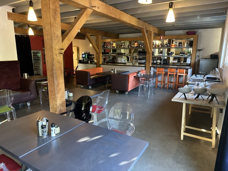 Modern café-restaurant interieur met houten balken, donkere tafels, banken, transparante stoelen en een bar met oranje krukken.