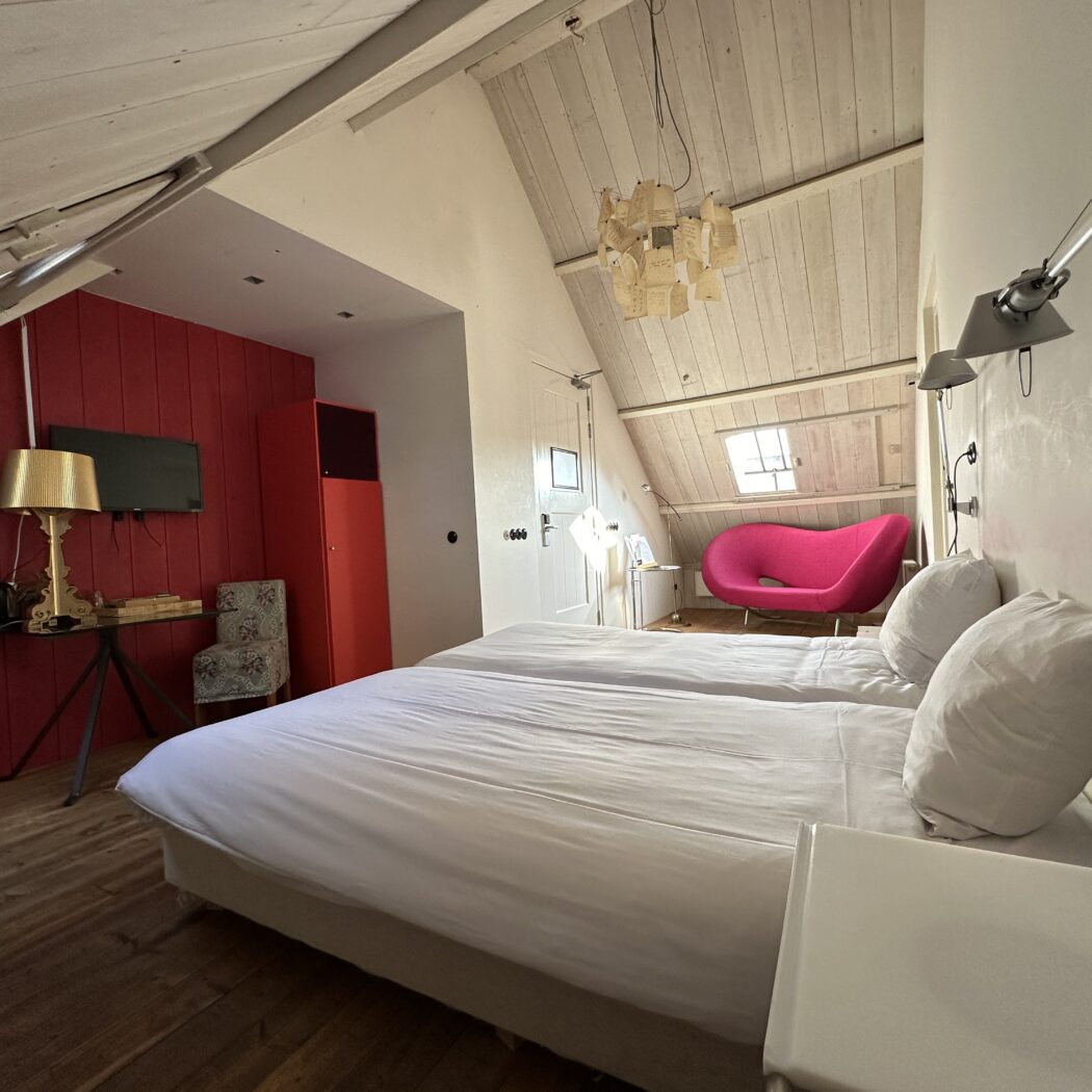 Lichte zolderkamer met twee witte bedden, rode accentmuur en fuchsia designstoel onder opvallende kroonluchter.