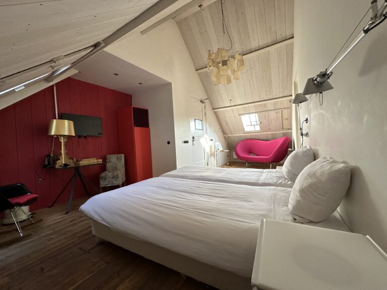 Lichte zolderkamer met twee witte bedden, rode accentmuur en fuchsia designstoel onder opvallende kroonluchter.