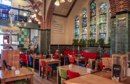 Sfeervol restaurantinterieur met gebrandschilderde ramen, groene tegels en eclectische verlichting boven kleurrijke zitjes.