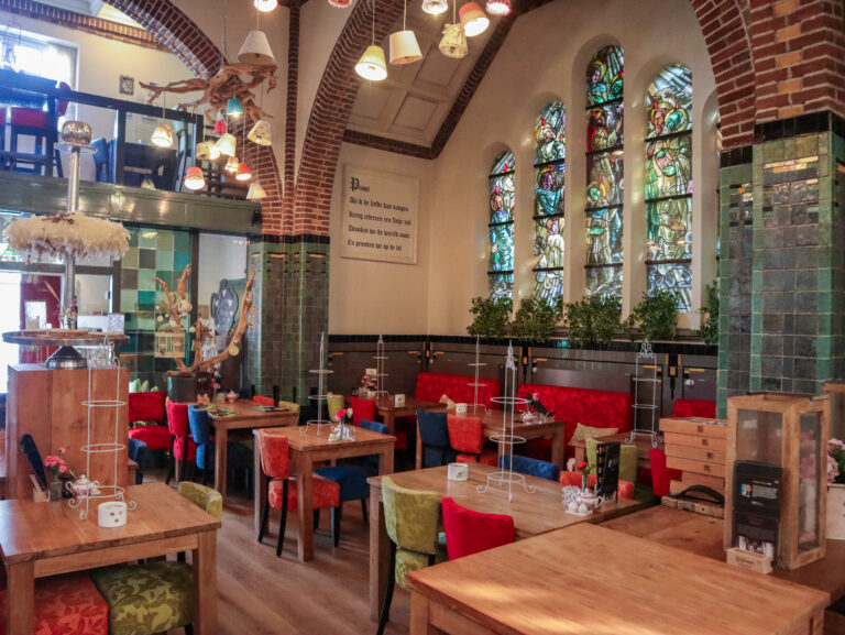 Sfeervol restaurantinterieur met gebrandschilderde ramen, groene tegels en eclectische verlichting boven kleurrijke zitjes.