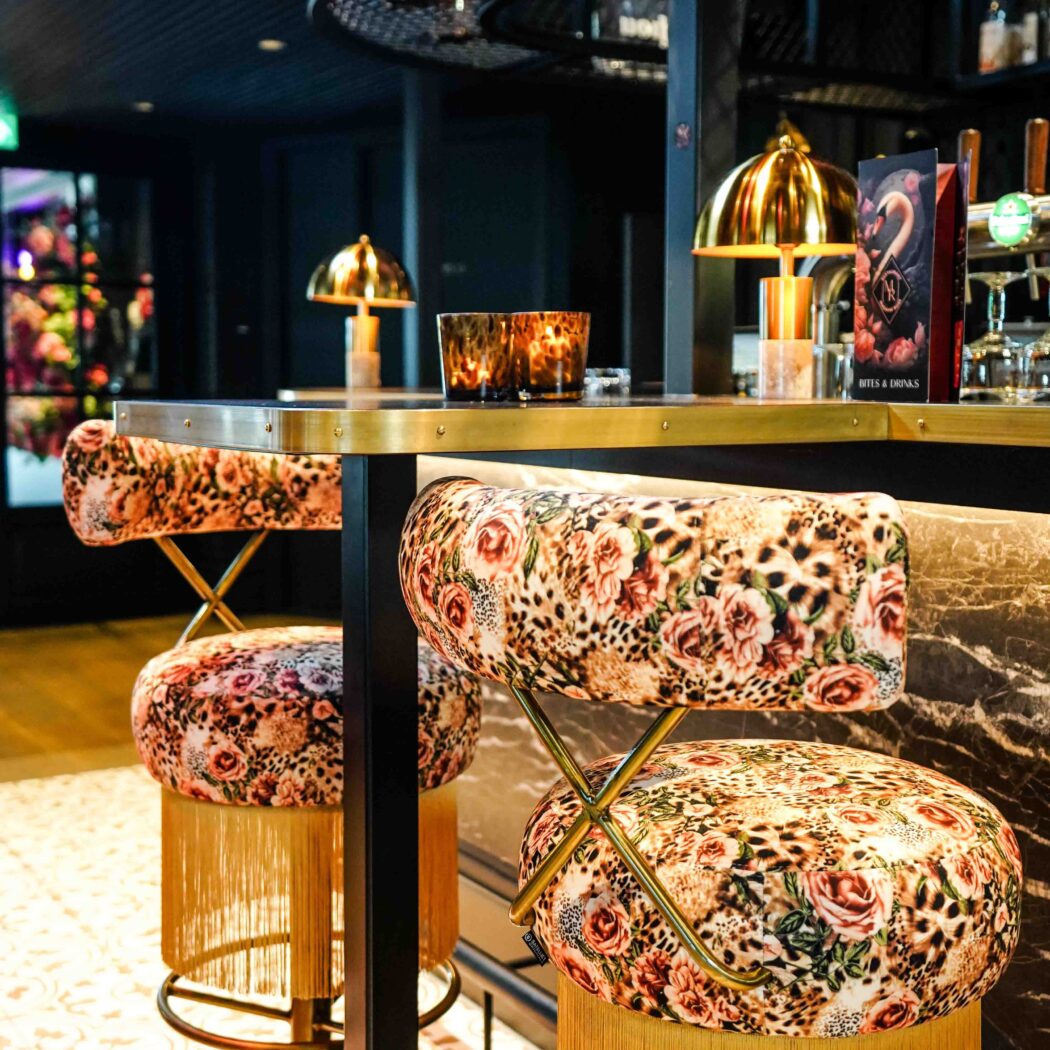 Twee barstoelen met bloemen- en luipaardprint en gouden franjes aan een bar met Bites & Drinks menu.