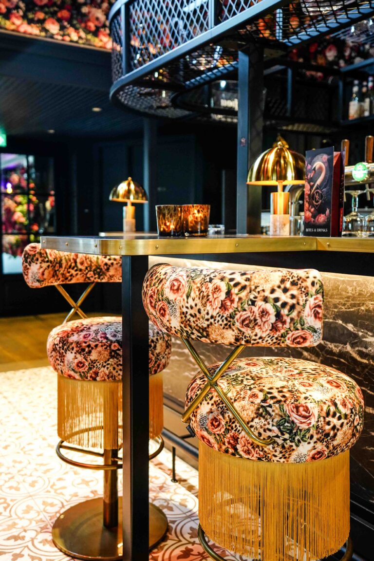 Twee barstoelen met bloemen- en luipaardprint en gouden franjes aan een bar met Bites & Drinks menu.
