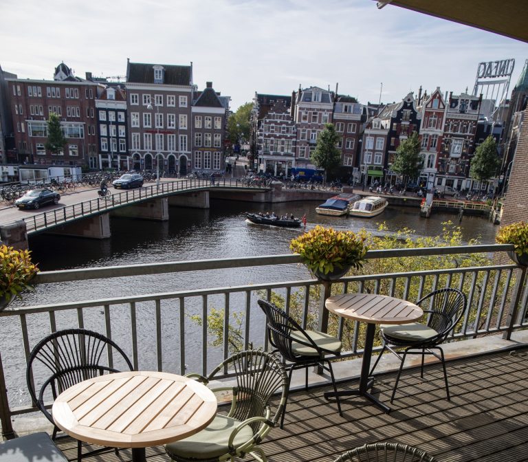 Zonnig balkon met houten tafels kijkt uit op een Amsterdamse gracht met boten, een brug en historische panden.