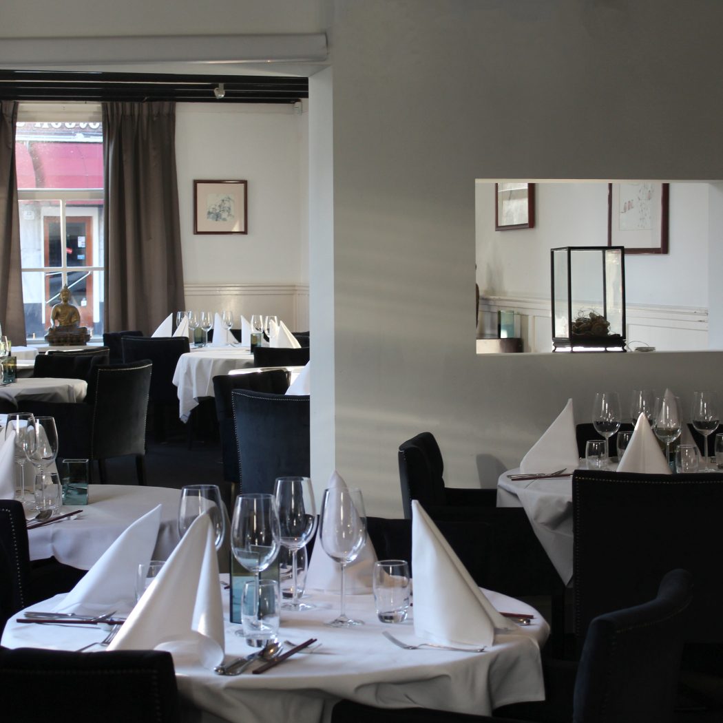 Interieur van een restaurant met gedekte tafels, donkere stoelen en een Boeddhabeeldje bij het raam.