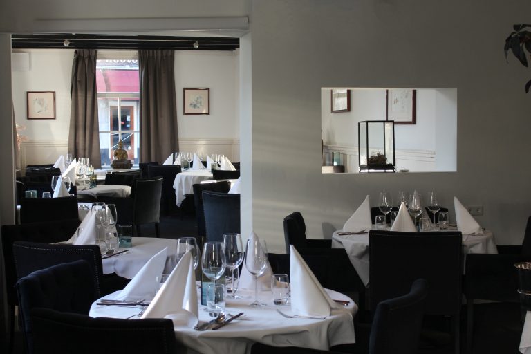Interieur van een restaurant met gedekte tafels, donkere stoelen en een Boeddhabeeldje bij het raam.