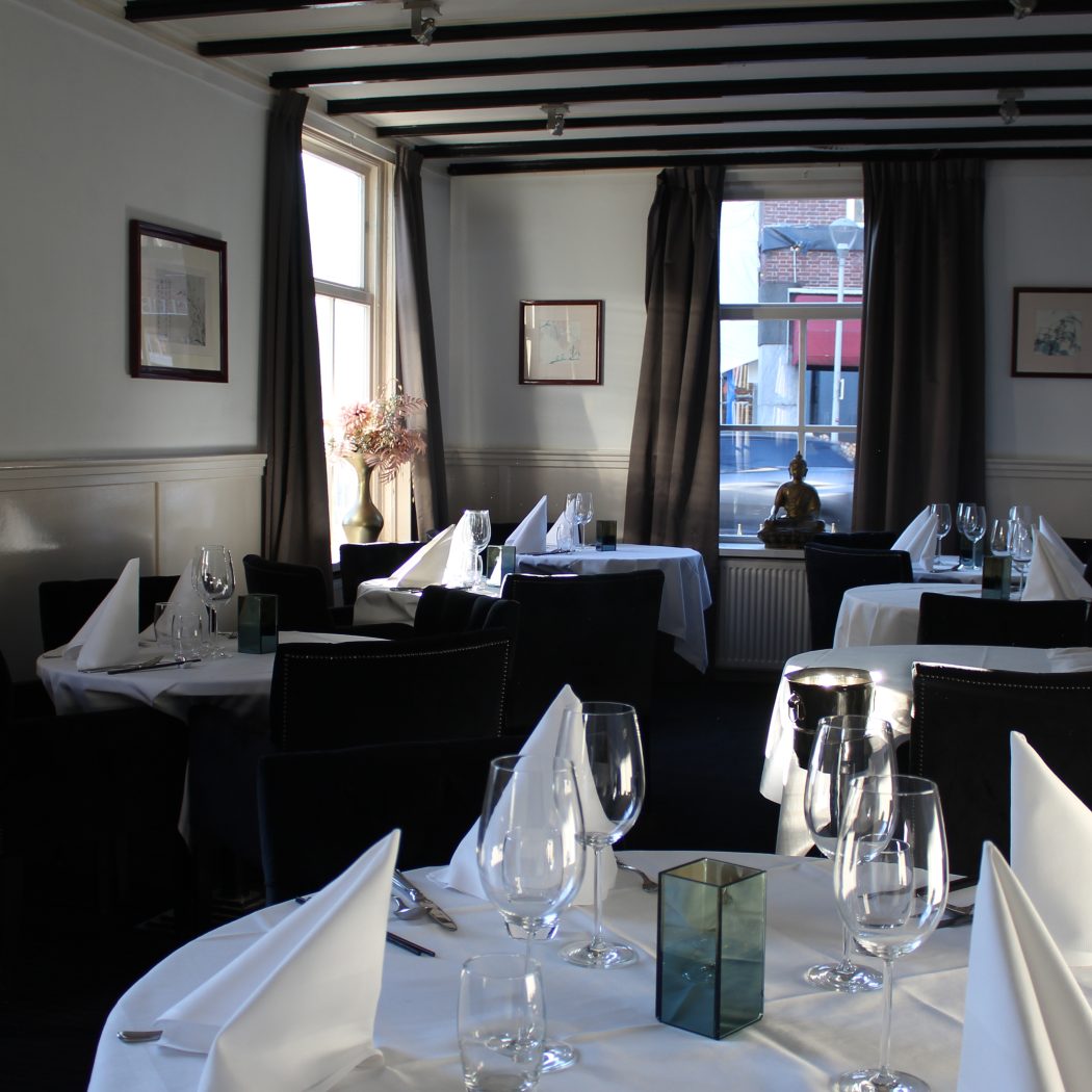 Elegant restaurant met gedekte tafels, witte linnen, glazen, bestek en zwarte stoelen.