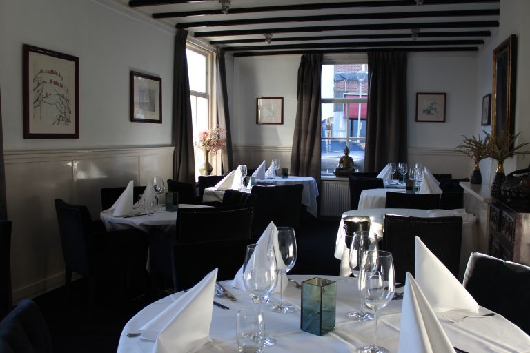 Elegant restaurant met gedekte tafels, witte linnen, glazen, bestek en zwarte stoelen.