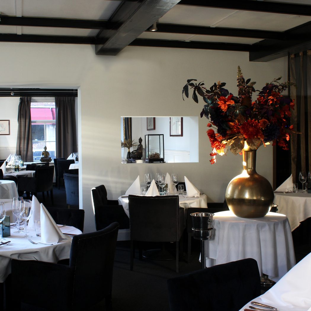 Luxe restaurant met wit gedekte tafels, zwarte stoelen, een gouden vaas met bloemen en donkere plafondbalken.