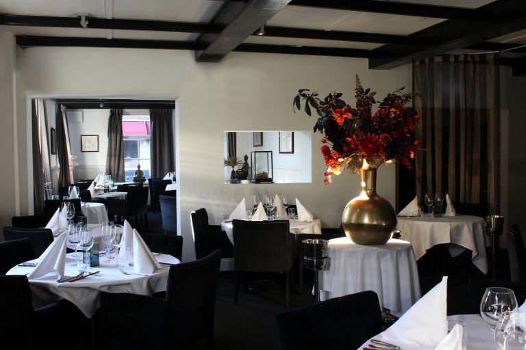 Luxe restaurant met wit gedekte tafels, zwarte stoelen, een gouden vaas met bloemen en donkere plafondbalken.