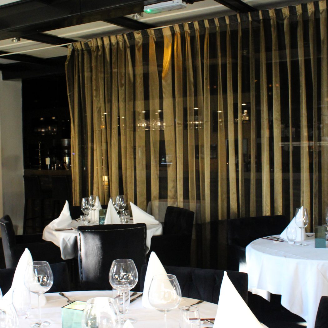 Sfeervol restaurant met gedekte tafels, donkere stoelen en opvallende goudbruin-zwarte gestreepte gordijnen.