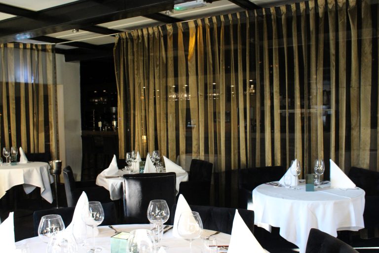 Sfeervol restaurant met gedekte tafels, donkere stoelen en opvallende goudbruin-zwarte gestreepte gordijnen.