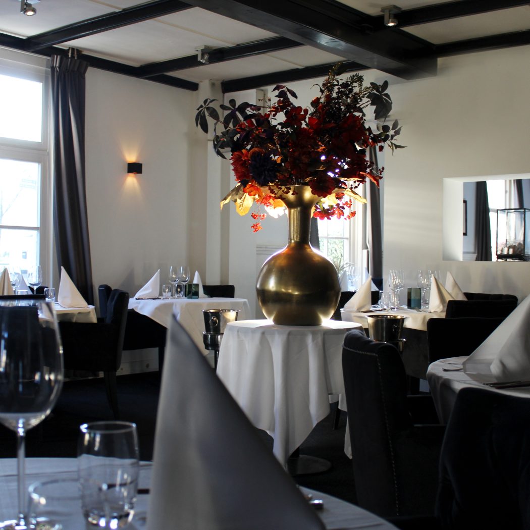 Ruime restaurantzaal met gedekte tafels, een grote gouden vaas met rode bloemen en donkere stoelen bij hoge ramen.