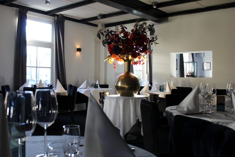 Ruime restaurantzaal met gedekte tafels, een grote gouden vaas met rode bloemen en donkere stoelen bij hoge ramen.