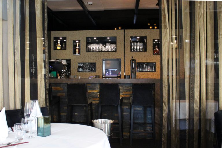 Moderne restaurantbar met flessen, glazen in verlichte nissen, drie zwarte barkrukken en biertap.