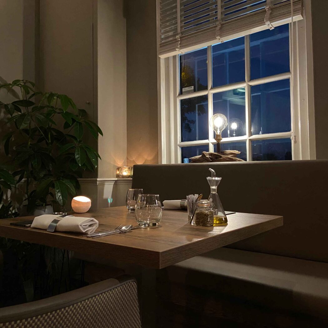 Gedekte houten restauranttafel met kaarslicht, olieflesje en plant, naast een raam met lamp in de avond.