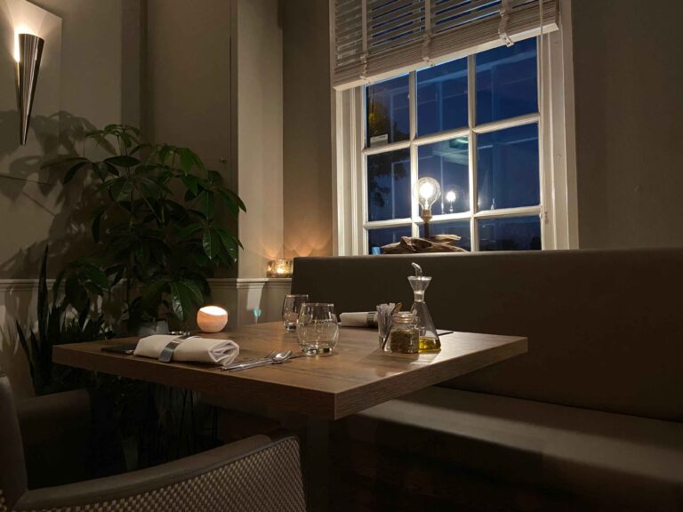 Gedekte houten restauranttafel met kaarslicht, olieflesje en plant, naast een raam met lamp in de avond.
