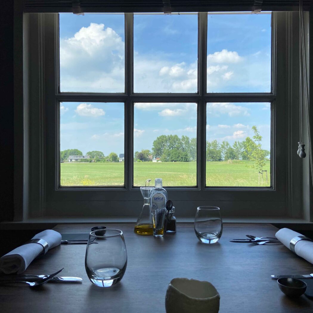 Gedekte houten tafel met twee plaatsen bij een raam dat uitkijkt op een zonnig groen veld met bomen en blauwe lucht.