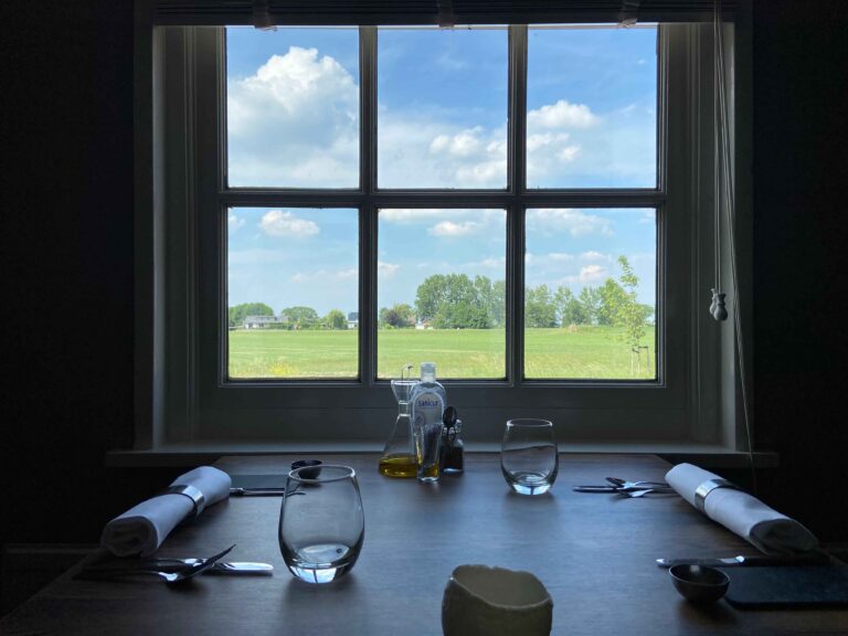 Gedekte houten tafel met twee plaatsen bij een raam dat uitkijkt op een zonnig groen veld met bomen en blauwe lucht.