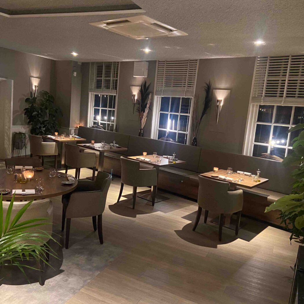 Sfeervol restaurantinterieur met gedekte tafels, comfortabele banken, planten en gedimd licht.