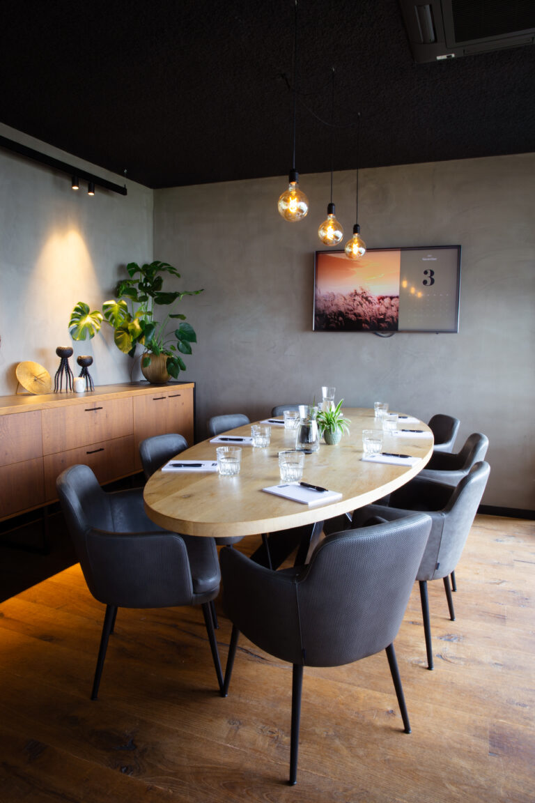 Moderne vergaderruimte met ovale houten tafel, donkergrijze stoelen, scherm met kalender en industriële lampen.