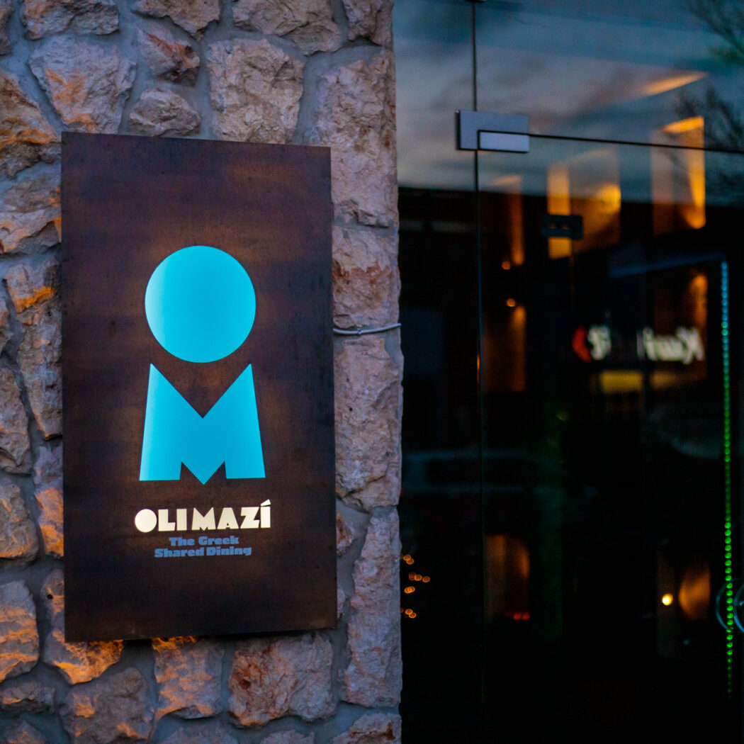 Verlicht Olimazí restaurantbord met blauw logo en The Greek Shared Dining op stenen muur, naast glazen deur.
