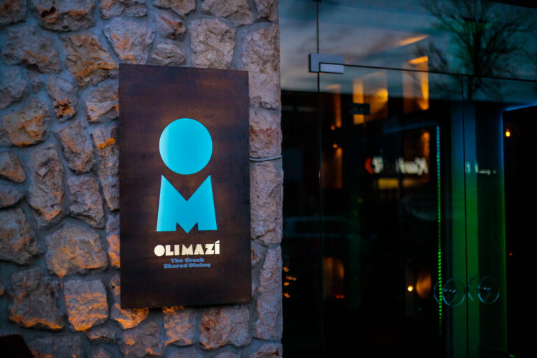 Verlicht Olimazí restaurantbord met blauw logo en The Greek Shared Dining op stenen muur, naast glazen deur.