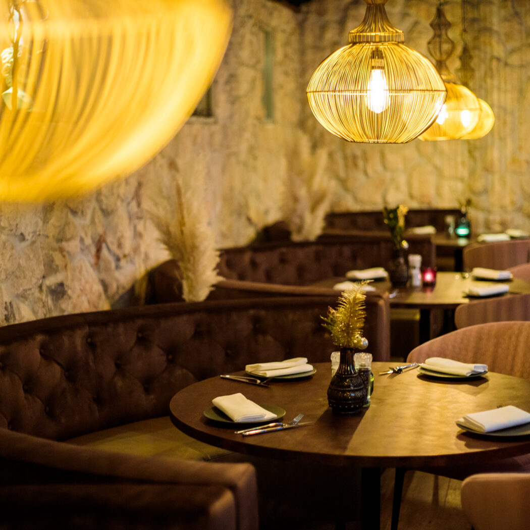 Sfeervol restaurantinterieur met ronde houten tafels, donkerbruine banken en gouden lampen tegen een stenen muur.