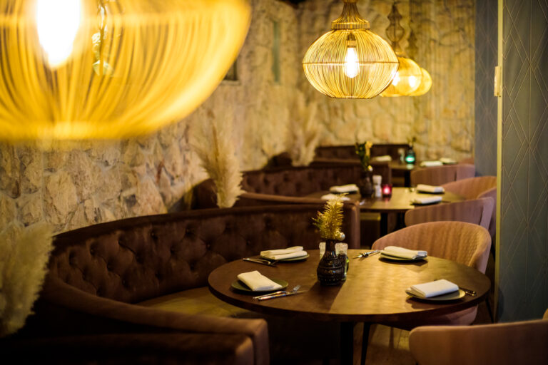Sfeervol restaurantinterieur met ronde houten tafels, donkerbruine banken en gouden lampen tegen een stenen muur.