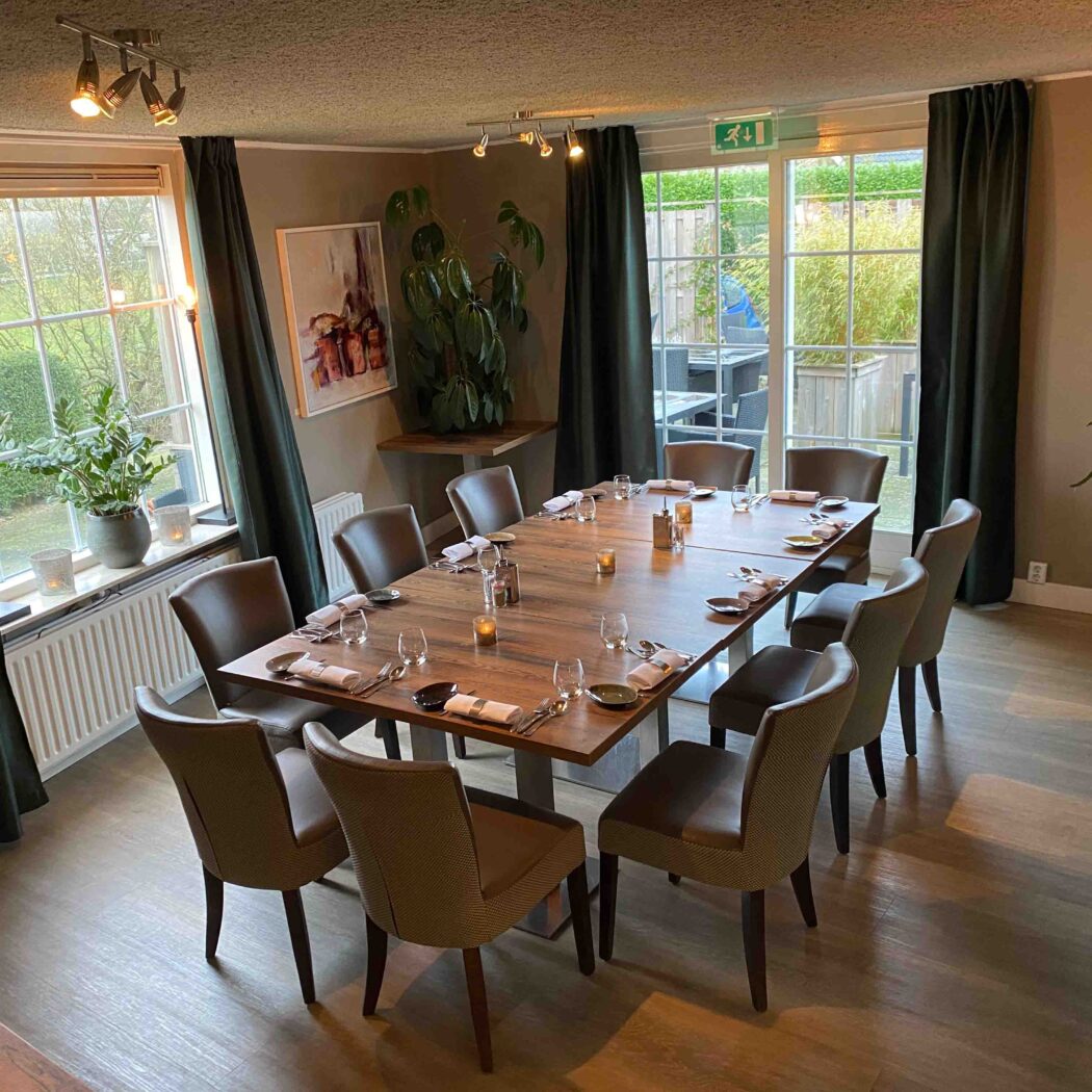 Sfeervolle privé eetruimte met lange houten tafel, gedekt voor tien personen, bij grote ramen.