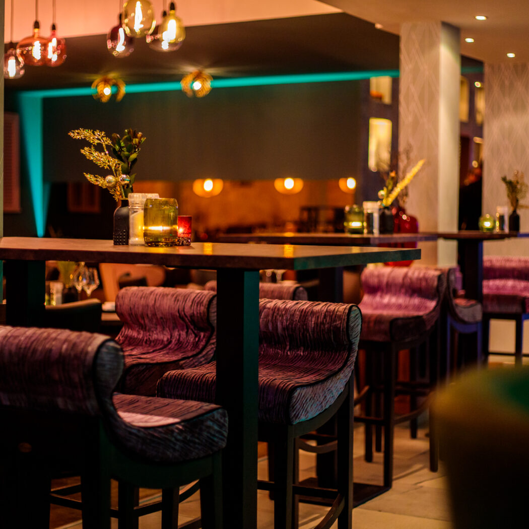 Sfeervol restaurantinterieur met hoge houten tafels, paarse barkrukken en warme kaarsverlichting.