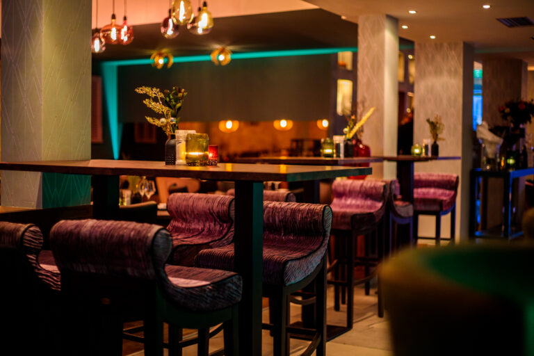Sfeervol restaurantinterieur met hoge houten tafels, paarse barkrukken en warme kaarsverlichting.