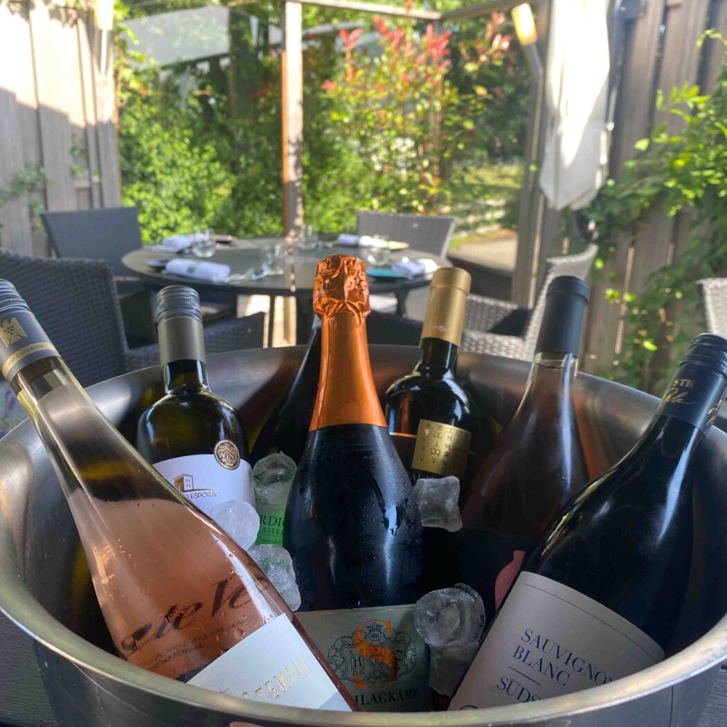 Zilveren wijnkoeler met flessen rosé (Enteza), witte wijn (Sauvignon Blanc) en champagne op ijs op een terras.