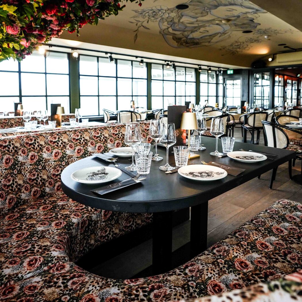 Stijlvol restaurant met bloemen- en luipaardprint banken, gedekte tafels, een bloeiende boom en geschilderd plafond.