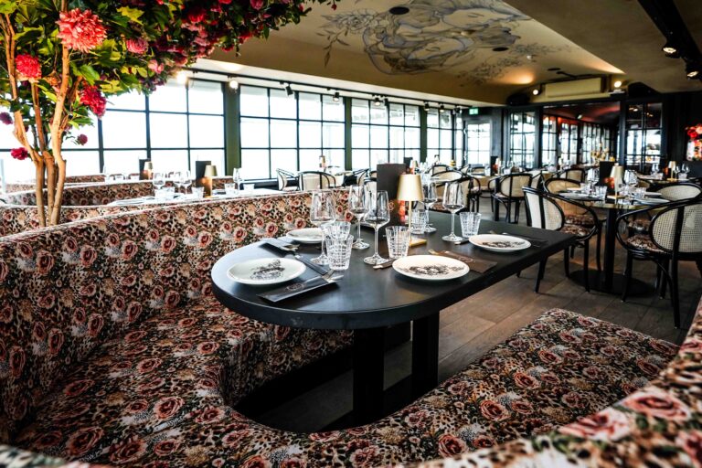 Stijlvol restaurant met bloemen- en luipaardprint banken, gedekte tafels, een bloeiende boom en geschilderd plafond.