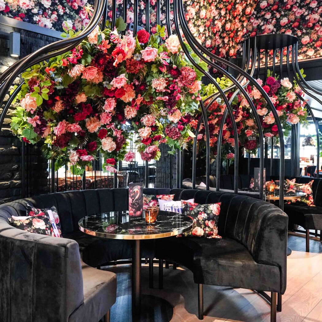 Chic restaurant met zwarte fluwelen banken, marmeren tafels en grote bloemdecoraties aan metalen bogen.