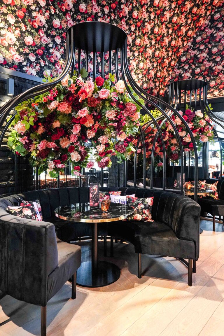 Chic restaurant met zwarte fluwelen banken, marmeren tafels en grote bloemdecoraties aan metalen bogen.