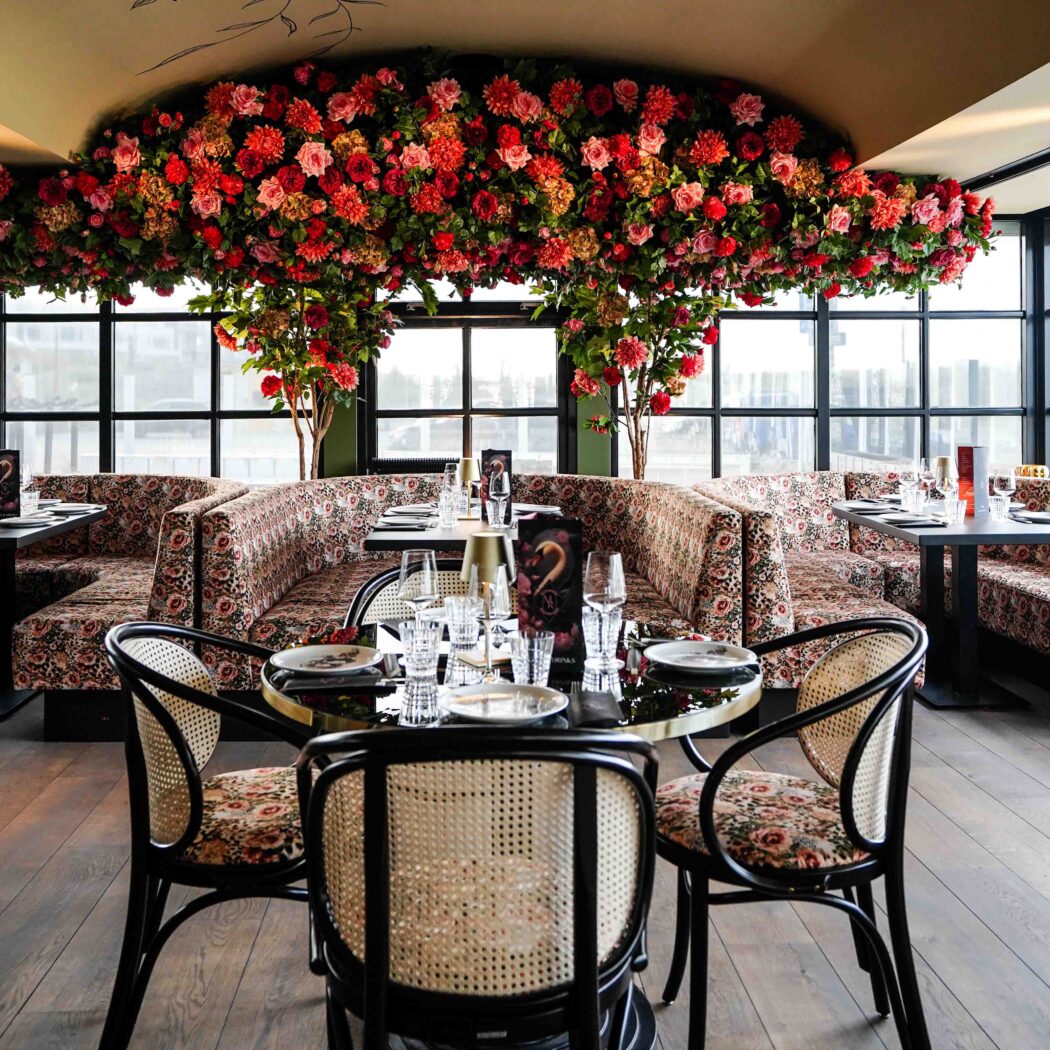 Sfeervol restaurant met rode en roze bloemen aan het plafond, gebloemde zitbanken, rieten stoelen en gedekte tafels bij ramen.