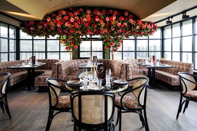 Sfeervol restaurant met rode en roze bloemen aan het plafond, gebloemde zitbanken, rieten stoelen en gedekte tafels bij ramen.