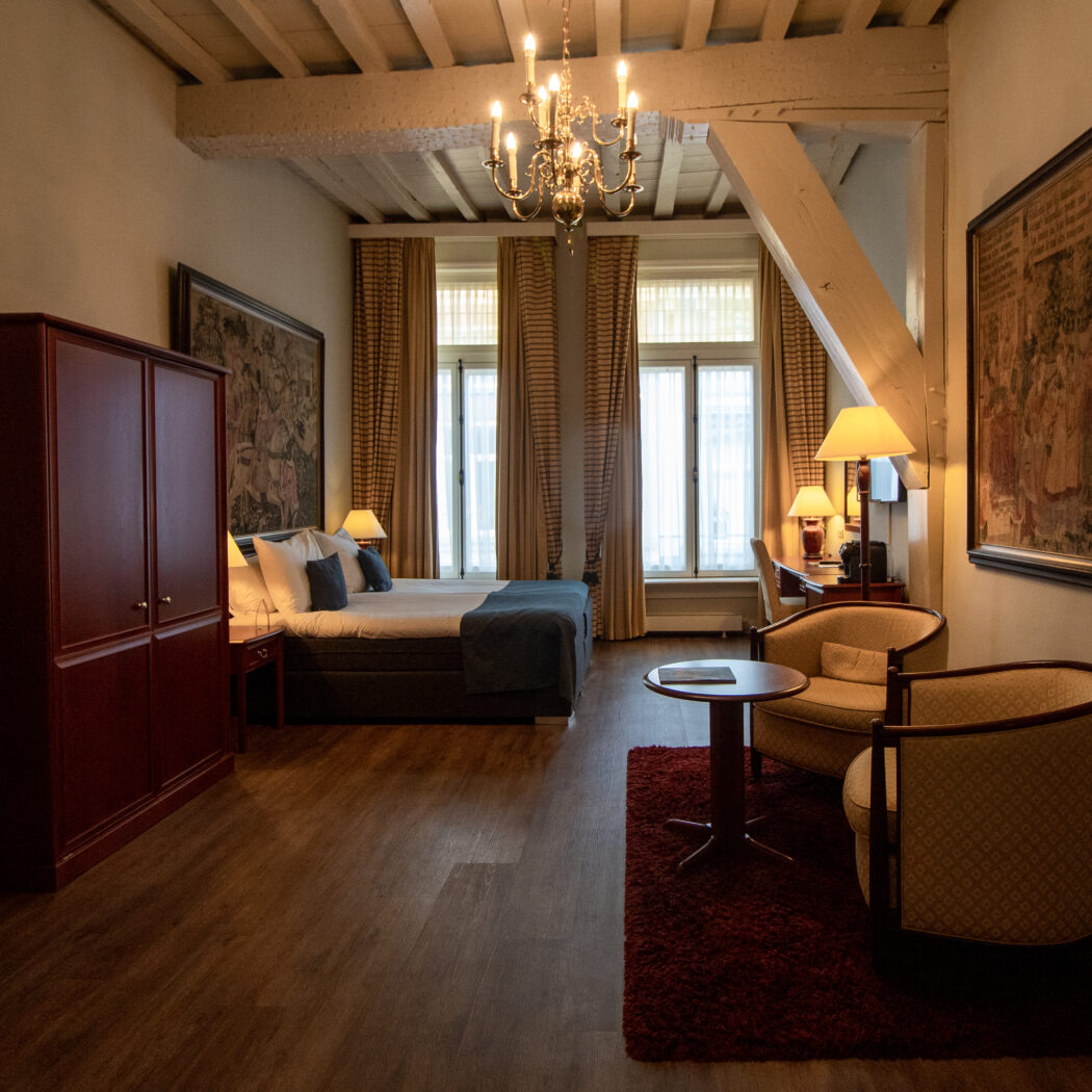Klassieke hotelkamer met kroonluchter, balkenplafond, een groot bed, rode kledingkast en zithoek.