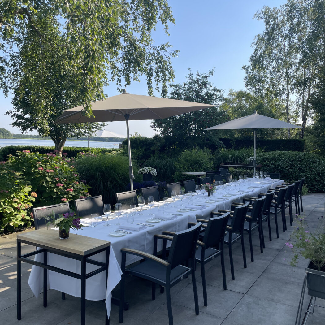 Lange, witgedekte buitentafel met bestek, glazen, donkere stoelen en parasols op een zonnig terras aan het water.
