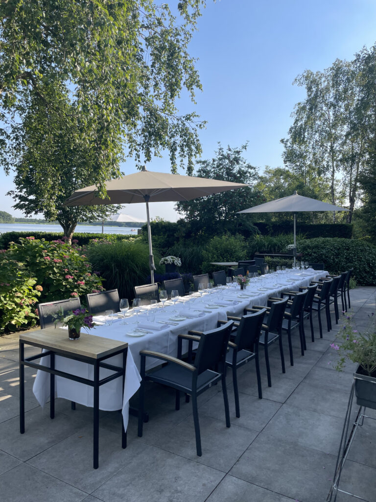 Lange, witgedekte buitentafel met bestek, glazen, donkere stoelen en parasols op een zonnig terras aan het water.