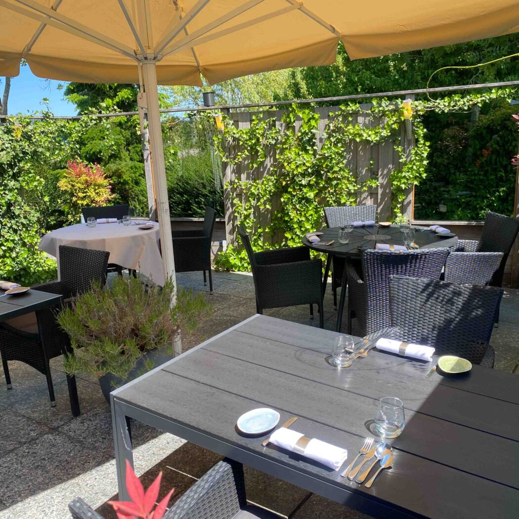 Uitnodigend buitenterras van een restaurant met gedekte tafels en donkere stoelen onder beigekleurige parasols, tussen weelderig groen.