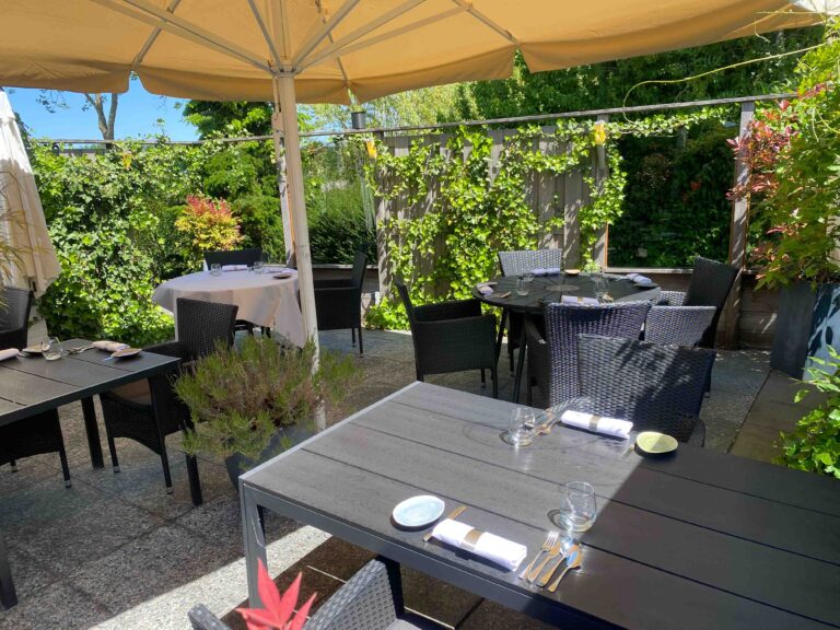 Uitnodigend buitenterras van een restaurant met gedekte tafels en donkere stoelen onder beigekleurige parasols, tussen weelderig groen.