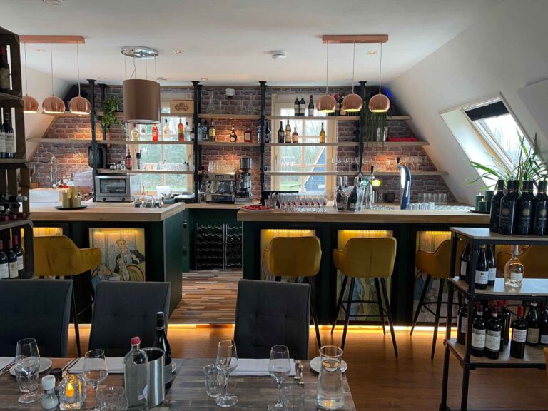 Sfeervol restaurantinterieur met houten bar, gele barstoelen, bakstenen muur en gevulde wijn- en drankrekken.