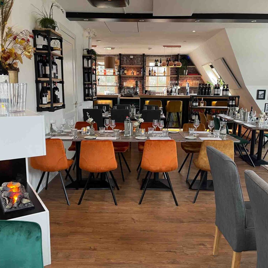 Sfeervol restaurantinterieur met lange tafels, oranje en groene stoelen, een bar en een decoratieve open haard.